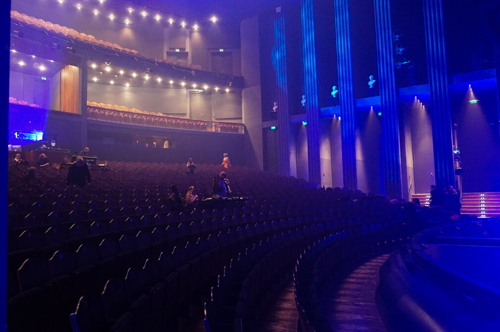Ein leerer Theaterraum mit vielen Sitzplätzen, die in sanften Bögen angeordnet sind. Einige Personen sind auf der Bühne und im Zuschauerbereich zu sehen, umgeben von bläulicher Beleuchtung. Die Atmosphäre wirkt ruhig und erwartungsvoll.