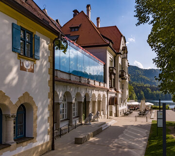 Das Museum der bayerischen Könige am Alpseeufer mit belebter Promenade und Blick auf die Berge.
