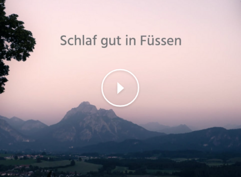 Das Bild zeigt eine in Abendlicht getauchte Landschaft mit einer imposanten Bergkulisse im Hintergrund. Der Himmel leuchtet in Lila- und Blautönen des letzten Sonnenlichts. Im Vordergrund rahmen zwei große Laubbäume das Bild links und rechts ein.