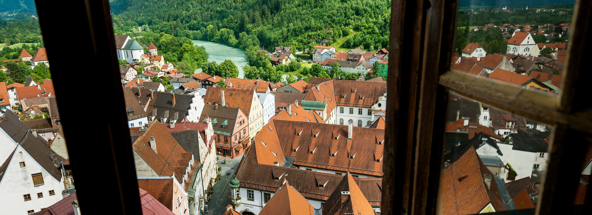 Das Foto zeigt den Blick aus einem Fenster des Hohen Schlosses, das hoch über der Stadt Füssen thront. Man sieht die Dächer der Altstadt von oben. Man kann sogar einen Blick in eine Gasse der Altstadt werfen. Im Hintergrund erheben sich die Berge. Davor fließt der türkise Lech. Es ist Frühling. Das Laub der Bäume auf dem Bergwald ist hellgrün. Der Himmel ist wolkenverhangen.  