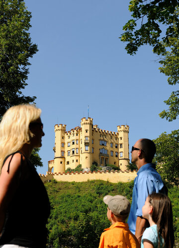 Das Bild zeigt eine friedliche Szene einer Familie bestehend aus einer Frau, einem Mann und zwei Kindern, die vor einem Schloss stehen. Die Frau, in einem schwarzen Oberteil gekleidet, steht links im Bild. Sie schaut in Richtung rechts, ihr Blick folgt dem Mann und den zwei Kindern. Der Mann trägt ein blaues Hemd und steht in der Mitte des Bildes, flankiert von den beiden Kindern, das eine in einem orangefarbenen Hemd und das andere in einem blauen Hemd. Das Schloss, das farblich in einem leuchtenden Gelb gestaltet ist, steht majestätisch im Hintergrund. Die Familie scheint einen Ausflug zu genießen und die Ansichten des Schlosses zu bewundern.