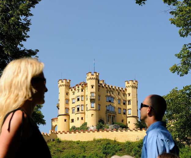 Das Bild zeigt eine friedliche Szene einer Familie bestehend aus einer Frau, einem Mann und zwei Kindern, die vor einem Schloss stehen. Die Frau, in einem schwarzen Oberteil gekleidet, steht links im Bild. Sie schaut in Richtung rechts, ihr Blick folgt dem Mann und den zwei Kindern. Der Mann trägt ein blaues Hemd und steht in der Mitte des Bildes, flankiert von den beiden Kindern, das eine in einem orangefarbenen Hemd und das andere in einem blauen Hemd. Das Schloss, das farblich in einem leuchtenden Gelb gestaltet ist, steht majestätisch im Hintergrund. Die Familie scheint einen Ausflug zu genießen und die Ansichten des Schlosses zu bewundern.