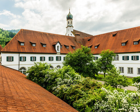 Das Bild zeigt das Franziskanerkloster in Füssen mit seinem markanten roten Ziegeldach und weißen Fassade. Üppige Bäume umgeben das Kloster.