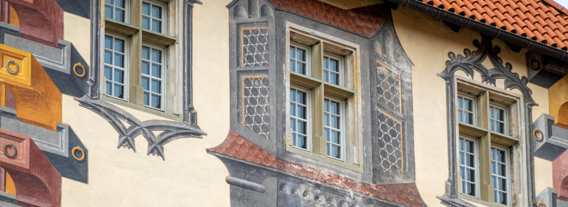 Das Bild zeigt einen Detail eines mit Illusionsmalerei verziertes Gebäude. Drei Fenster stehen im Fokus. Ein kleiner Teil des roten Dachs ist zu sehen. 
