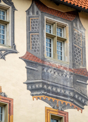 Das Bild zeigt einen Detail eines mit Illusionsmalerei verziertes Gebäude. Drei Fenster stehen im Fokus. Ein kleiner Teil des roten Dachs ist zu sehen. 