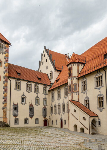 Das Bild zeigt den Innenhof des Hohen Schlosses in Füssen, der durch seine Illusionsmalerei an den Fassaden beeindruckt. Das Schlossgebäude hat rote Türen und ein Ziegeldach.
