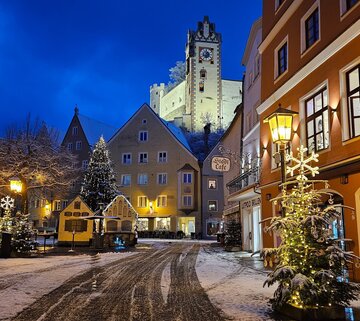 Ein verschneiter Blick auf eine malerische Füssener Altstadt. Die warmen Lichter der Häuser und Laternen erzeugen eine gemütliche und festliche Atmosphäre.