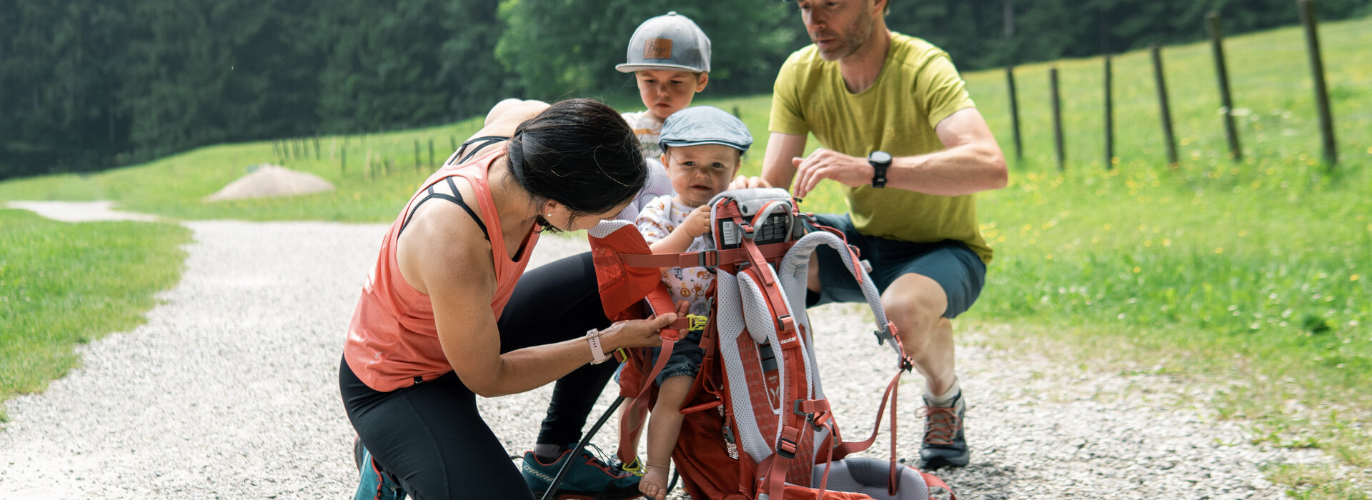 Eine Frau befestigt ein Kleinkind in einem Wanderrucksack, während ein Mann und ein weiteres Kind zuschauen. Die Szene spielt sich auf einem Kiesweg in einer grünen Landschaft ab, umgeben von Bäumen und Wiesen.