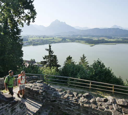 Das Bild zeigt eine friedvolle Szene von zwei Menschen, einem Mann und einer Frau, die in den alten Mauern einer verfallenen Burg herumgehen. Die Ruine befindet sich auf einer Anhöhe über dem Hopfensee und ist von einer lichten Berglandschaft und einem dichten Wald umgeben. Die Bergkette, majestätisch und hoch, bildet eine beeindruckende Kulisse für diese Szenerie. Der Himmel ist ein helles Blau, was zu der allgemeinen Ruhe des Bildes beiträgt. Die beiden Menschen, in legerer Kleidung, scheinen einen gemütlichen Spaziergang am Ufer zu genießen. Das Bild vermittelt ein Gefühl von Frieden und Ruhe und ist somit ein faszinierender Anblick.