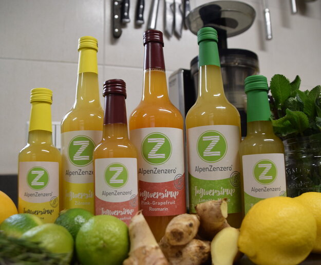 Das Bild zeigt verschiedene Sorten und Flaschengrößen des AlpenZenzero Sirups und die Zutaten wie Limette, Zitrone und Ingwer