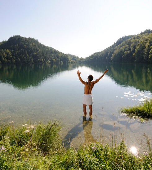 Das Bild zeigt einen Mann, der sich nur ein weißen Handtuch um die Hüften gebunden hat. Er hat dem Betrachter den Rücken zugedreht, steht im knöcheltiefen Wasser eines Sees und streckt die Arme nach oben. Der See ist umringt von grünen Wäldern. Der Himmel ist blau. Die Sonne scheint.        