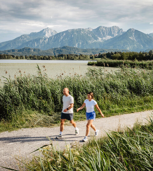 Das Bild zeigt eine Frau und einen Mann, die am Ufer des Hopfensees sportlich spazieren gehen