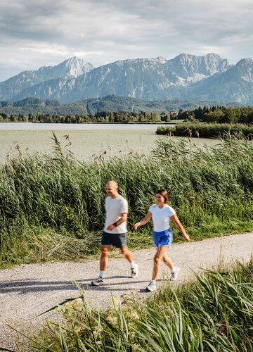 Das Bild zeigt einen Mann und eine Frau, die am Ufer des Hopfensees spazieren gehen.