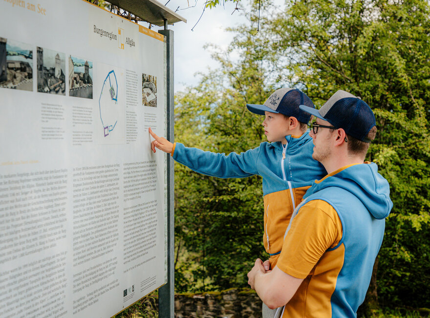 Das Bild zeigt einen Mann der einen Jungen auf dem Arm hält. Sie blicken gemeinsam auf eine Informationstafel.
