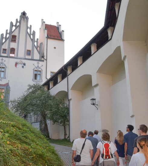 Das Bild zeigt eine Gruppe Menschen, die dem Betrachter weggewandt sind. Die Gruppe steht im Schlosshof des Hohen Schlosses und laufen in Richtung des Hauptgebäudes. Die Abendsonne taucht die Szene in ein warmes Licht. 