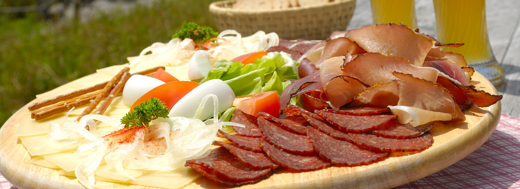 Eine Holzplatte mit einer Allgäuer Brotzeit bestehend aus Wurst, Käse und Brot, dazu zwei Biergläser, vor einer schönen Bergkulisse.
