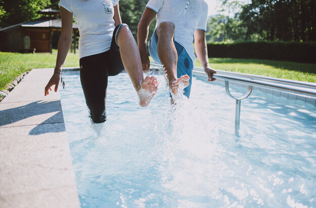 Das Bild zeigt zwei Personen, die in einem Kneipptretbecken stehen. Man sieht nicht ihre Gesichter. Beide Personen heben ein Bein aus dem Wasser, sodass die Wassertropfen um sie herum spritzen. im Hintergrund zeigt sich ein grüner Park mit satten Wiesen und belaubten Bäumen.