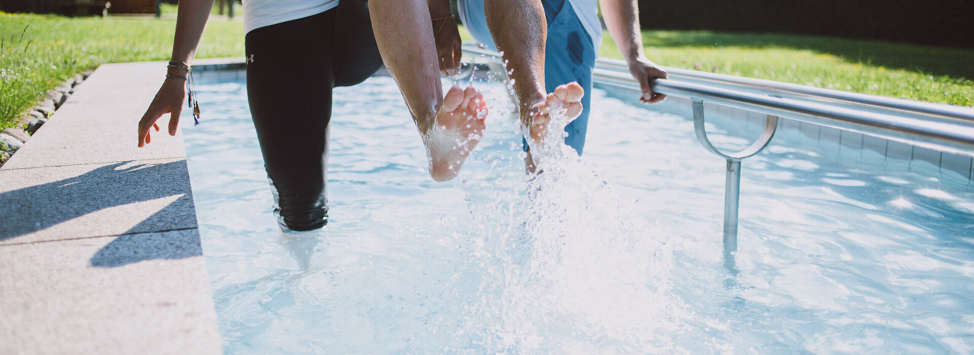 Das Bild zeigt zwei Personen, die in einem Kneipptretbecken stehen. Man sieht nicht ihre Gesichter. Beide Personen heben ein Bein aus dem Wasser, sodass die Wassertropfen um sie herum spritzen. im Hintergrund zeigt sich ein grüner Park mit satten Wiesen und belaubten Bäumen.