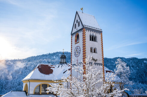 Dach einer barocken Kirche mit Kirchturm vor schneebedecktem Wald. Schnee auf Dächern und Ästen im Vordergrund unter blauem Himmel.