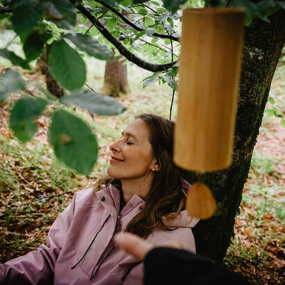 Eine Ferau lehnt an einem Baum und lauscht einem Windspiel