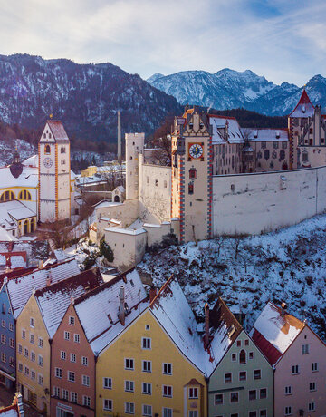 Das Bild fängt die atemberaubende Sicht auf das mittelalterliche Städtchen Füssen in Deutschland ein. Die Stadt liegt auf einem Hügel, von majestätischen Bergen umgeben. Die Architektur der Stadt ist durch bunte Gebäude geprägt, jedes mit einem einzigartigen Charme. Die Gebäude sind mit Schnee verziert, was eine Prise Winter in die Szene bringt