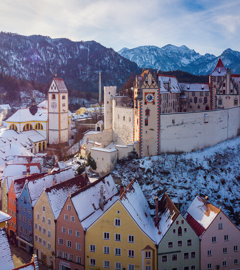 Das Bild fängt die atemberaubende Sicht auf das mittelalterliche Städtchen Füssen in Deutschland ein. Die Stadt liegt auf einem Hügel, von majestätischen Bergen umgeben. Die Architektur der Stadt ist durch bunte Gebäude geprägt, jedes mit einem einzigartigen Charme. Die Gebäude sind mit Schnee verziert, was eine Prise Winter in die Szene bringt