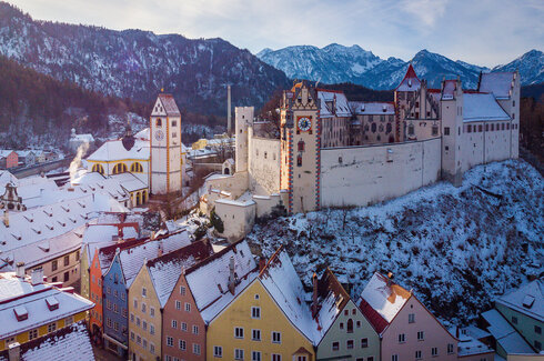 Das Bild fängt die atemberaubende Sicht auf das mittelalterliche Städtchen Füssen in Deutschland ein. Die Stadt liegt auf einem Hügel, von majestätischen Bergen umgeben. Die Architektur der Stadt ist durch bunte Gebäude geprägt, jedes mit einem einzigartigen Charme. Die Gebäude sind mit Schnee verziert, was eine Prise Winter in die Szene bringt