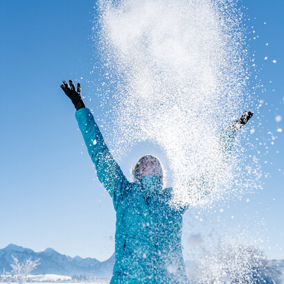 Das Bild zeigt eine Frau in blauer Outdoor-Jacke, die mit beiden Händen glitzernden Schnee in den blauen Himmel wirft.