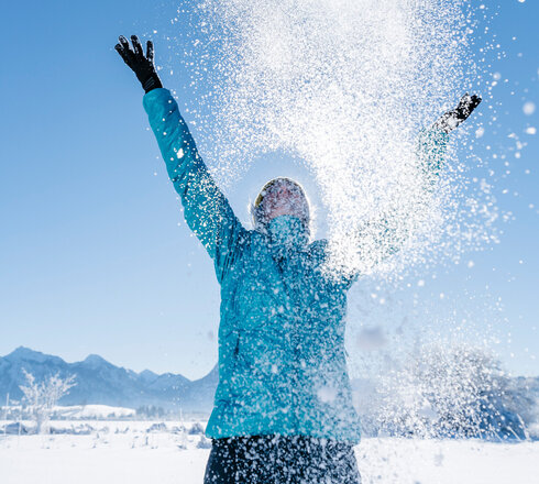 Das Bild zeigt eine Frau in blauer Outdoor-Jacke, die mit beiden Händen glitzernden Schnee in den blauen Himmel wirft.