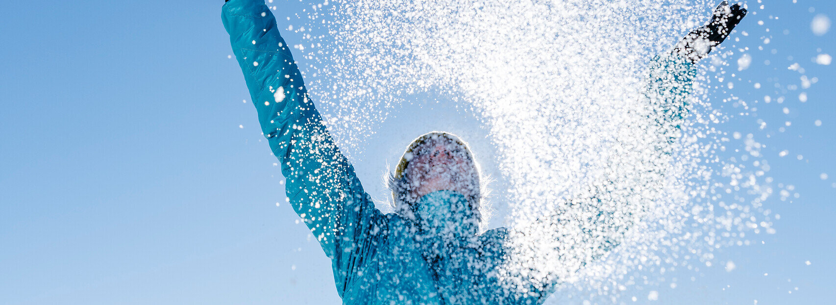 Das Bild zeigt eine Frau in blauer Outdoor-Jacke, die mit beiden Händen glitzernden Schnee in den blauen Himmel wirft.
