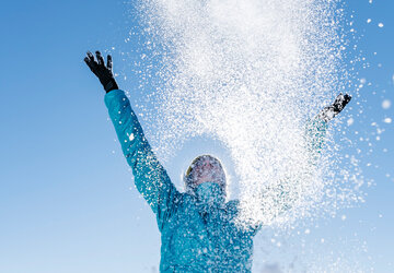 Das Bild zeigt eine Frau in blauer Outdoor-Jacke, die mit beiden Händen glitzernden Schnee in den blauen Himmel wirft.