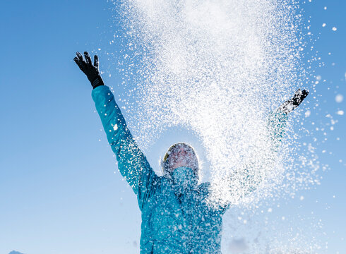 Das Bild zeigt eine Frau in blauer Outdoor-Jacke, die mit beiden Händen glitzernden Schnee in den blauen Himmel wirft.