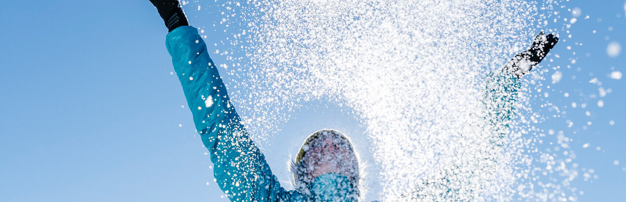 Das Bild zeigt eine Frau in blauer Outdoor-Jacke, die mit beiden Händen glitzernden Schnee in den blauen Himmel wirft.