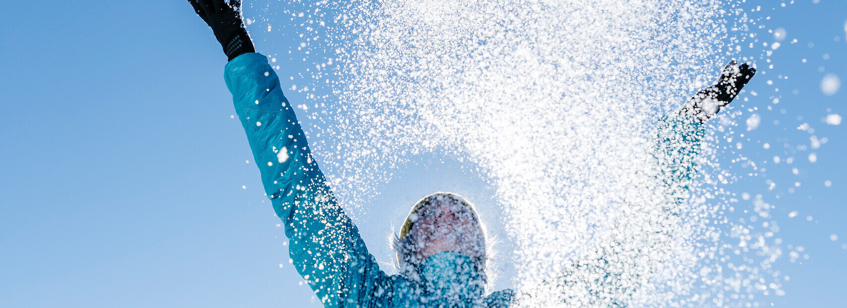 Das Bild zeigt eine Frau in blauer Outdoor-Jacke, die mit beiden Händen glitzernden Schnee in den blauen Himmel wirft.