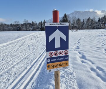 Das Bild zeigt ein Schild an einem Stab mit einem großen weißen Pfeil auf blauem Grund und weiteren Icon darauf abgebildet. Im Hintergrund zieht sich eine präparierte Langlaufloipe durch den Schnee und schneebedeckte Gipfel leuchten in der hellen Sonne.