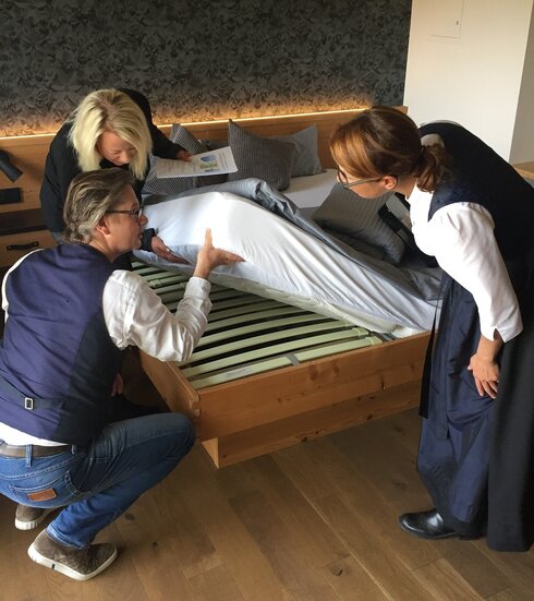 Das Bild zeigt drei Personen, die die Matratze eine Bettes hochheben und den darunterliegenden Lattenrost betrachten. Eine Frau trägt ein dunkles Dirndl. Eine Frau hat Papiere in der Hand. Ein Mann in Jeans, weißem Hemd und Weste ist in die Hocke gegangen.