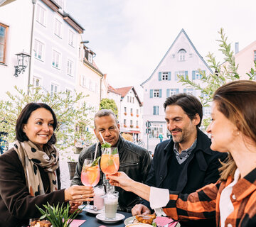 Eine Gruppe von Freunden sitzt an einem Tisch im Freien in einem Füssener Café und erhebt ihre Gläser zu einem fröhlichen Anstoß.