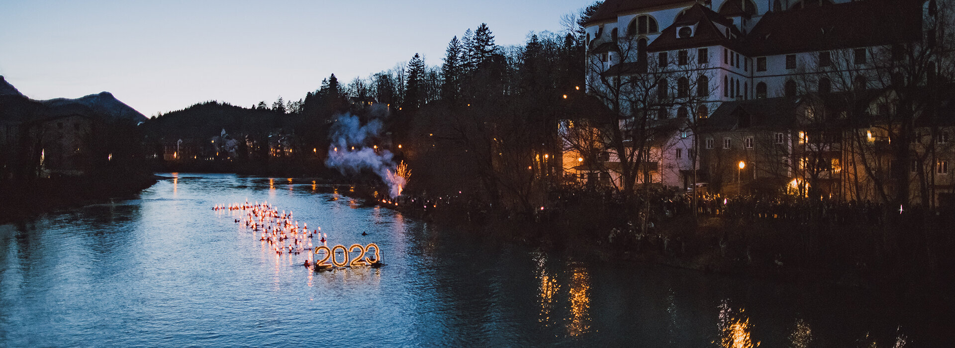 Das Bild zeigt eine Nachtszene am Fluss Lech. Auf dem Fluss schwimmen Menschen, in Neoprenanzügen gekleidet, mit Fackeln in den Händen. Sie schieben eine Plattform im Wasser vor sich her auf der die beleuchtete Zahl "2023" montiert ist. Lichter sind auch entlang des Flusses verteilt und schaffen eine wunderschöne und festliche Atmosphäre. Der rechte Teil des Bildes zeigt einen großen Gebäudekomplex mit einer Kuppel. Das Gebäude ist beleuchtet und trägt dazu bei, die Gesamtheit der Szene zu erhellen. Der Himmel darüber ist tiefblau. Das Bild wird aus der Ferne aufgenommen, was eine Panoramasicht des Flusses und des Gebäudes bietet. Die Gesamtzusammensetzung des Bildes erzeugt eine ruhige und feierliche Stimmung.