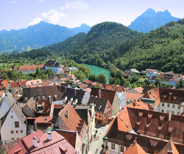 Das Foto zeigt Füssen von oben. Die verwinkelten roten Dächer der Altstadt mit Ihren bunten Fassaden reihen sich aneinander. Im Hintergrund erheben sich von grünem Bergwald bedeckte Berge. Davor fließt der türkisblaue Fluß Lech. 