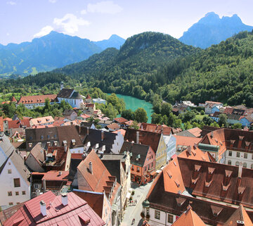 Das Foto zeigt Füssen von oben. Die verwinkelten roten Dächer der Altstadt mit Ihren bunten Fassaden reihen sich aneinander. Im Hintergrund erheben sich von grünem Bergwald bedeckte Berge. Davor fließt der türkisblaue Fluß Lech.