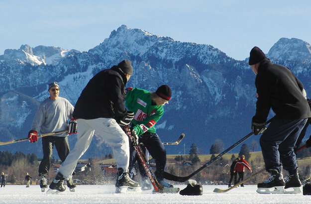 Das Bild zeigt mehrere Menschen beim Eishockey-Spielen auf einem zugefroreren See mit Blick auf die umliegenden Berge