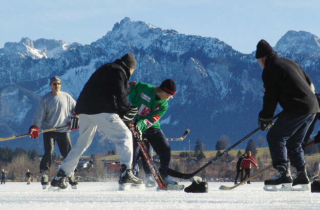 Das Bild zeigt mehrere Menschen beim Eishockey-Spielen auf einem zugefroreren See mit Blick auf die umliegenden Berge