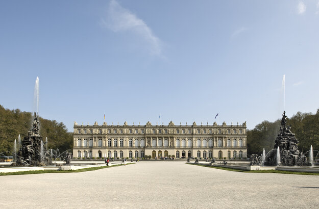 Das Bild zeigt die Aussenansicht des Schloss Herrenchiemsee. Vor dem Schloss sind rechts und links je ein Springbrunnen mit einer hohen Wasserfontäne zu sehen.
